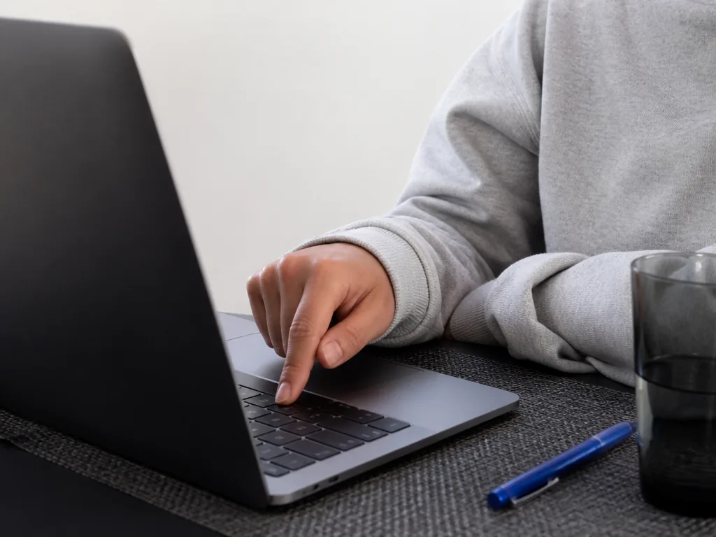 Photorealistic editorial photograph of a student's hand pressing a matte metal keyboard key on a blank laptop, desk with